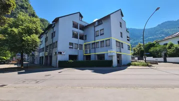 Mehrstöckiges Gebäude mit hellgrauer Fassade und vielen Fenstern, umgeben von Bäumen und Bergen unter blauem Himmel.