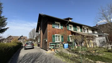 Traditionelles Holzhaus mit grünen Fensterläden und gepflasterter Auffahrt unter blauem Himmel.