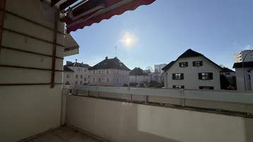 Helles Balkon mit Fliesenboden und Blick auf umliegende Gebäude unter blauem Himmel.