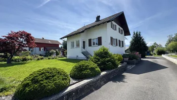 Weißes Einfamilienhaus mit Satteldach, gepflegtem Vorgarten und Zufahrt unter blauem Himmel.