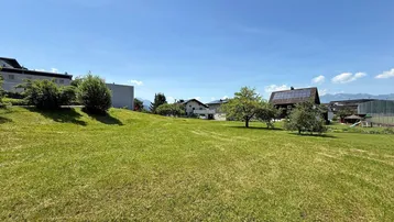 Weitläufige grüne Wiese mit umliegenden Häusern und Bäumen unter blauem Himmel.