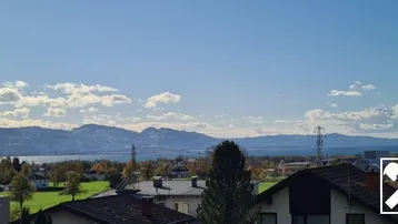 Panoramablick über einen See mit Bergen im Hintergrund und Dächern im Vordergrund unter blauem Himmel.