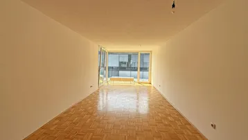 Helles, leeres Wohnzimmer mit Parkettboden und Zugang zu einem Balkon oder einer Terrasse.