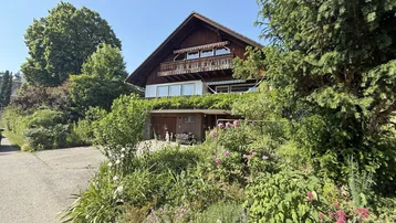Traditionelles Holzhaus mit Balkon und üppigem Garten, umgeben von viel Grün und Bäumen.
