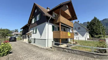 Charmantes Haus mit traditioneller Holzfassade, Balkonen und gepflasterter Auffahrt unter blauem Himmel.
