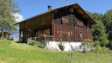 Traditionelles Holzhaus in den Bergen mit grüner Wiese und blauem Himmel an einem sonnigen Tag.