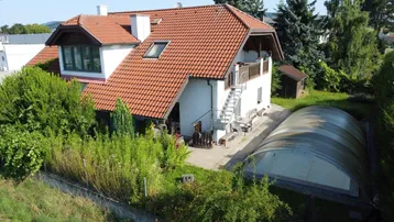 Luftaufnahme eines Einfamilienhauses mit rotem Ziegeldach, großem Garten und überdachtem Poolbereich.