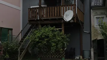 Hinteransicht des Hauses mit überdachtem Holzbalkon, Außentreppe und kleinem Rasenbereich.
