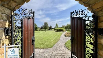 Prunkvolles schmiedeeisernes Tor öffnet sich zu einem weitläufigen, gepflegten Garten mit Steinweg.