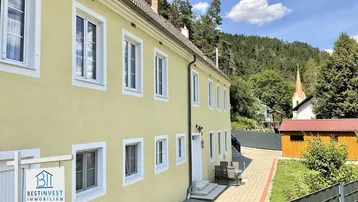 Gelbe Fassade eines Gebäudes mit weißen Fenstern, umgeben von viel Grün und einem Kirchturm in der Ferne.