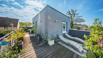 Geräumige Dachterrasse mit Holzbelag, Liegestühlen und Pflanzen unter blauem Himmel.