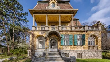 Historische Villa mit gelber Fassade, grünen Fensterläden und großem Balkon, umgeben von altem Baumbestand.