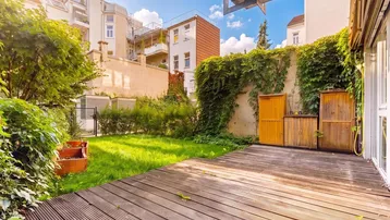 Große Holzterrasse grenzt an einen gepflegten Rasenbereich mit üppiger Bepflanzung und hohen Mauern.