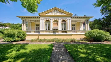 Klassische Villa mit eleganter Fassade, Säulen und gepflegtem Vorgarten unter blauem Himmel.