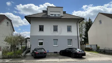 Vorderansicht eines Mehrfamilienhauses mit zwei geparkten Autos und einem kleinen Vorgarten.