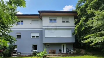 Mehrstöckiges Wohngebäude mit grauer und weißer Fassade, umgeben von üppigem Grün und einem kleinen Gartenbereich mit Sitzgelegenheiten.