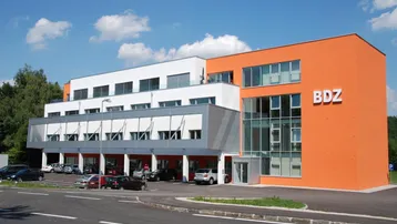 Modernes Bürogebäude mit orangefarbener Fassade, großen Fenstern und Parkplätzen davor unter blauem Himmel.