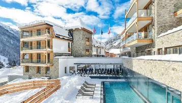 Modernes Gebäude in alpiner Winterlandschaft mit Außenpool und Liegestühlen unter blauem Himmel.