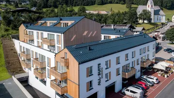Modernes Apartmentgebäude mit Holzelementen und Balkonen, umgeben von grünen Hügeln und einer Kirche.