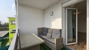 Sonniger Balkon mit bequemen Rattanmöbeln, Kunstrasen und Blick auf die grüne Umgebung.