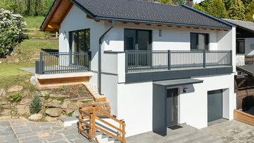 Modernes Einfamilienhaus mit weißer Fassade, dunklem Dach und Balkon in grüner Umgebung.