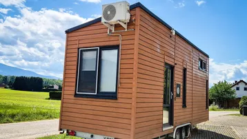 Modernes Tiny House mit Holzfassade und Klimaanlage, umgeben von grüner Landschaft.