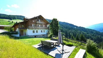 Traditionelles Holzhaus mit weißer Fassade, umgeben von grünen Wiesen und Wäldern, mit einer Terrasse im Vordergrund.