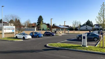 Ein moderner E-Ladepark mit mehreren Ladestationen für Elektrofahrzeuge unter blauem Himmel und umgeben von Grünflächen.