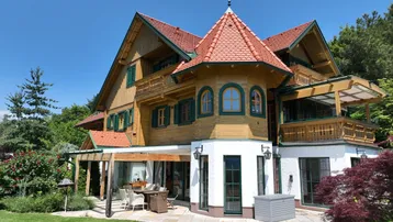 Traditionelles Holzhaus mit rotem Ziegeldach und Turm, davor eine möblierte Terrasse und Rasenfläche.