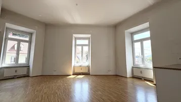 Helles, leeres Wohnzimmer mit Parkettboden und mehreren Fenstern, die viel Tageslicht hereinlassen.