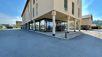 Modernes Gebäude mit großen Glasfronten im Erdgeschoss und weitläufigem Vorplatz unter blauem Himmel.