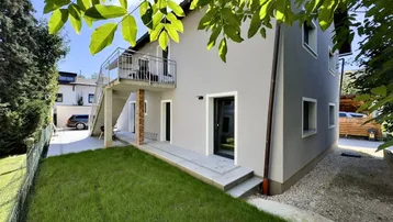 Modernes Gebäude mit grauem Putz, kleinem Rasenbereich und Außentreppe zum Balkon.