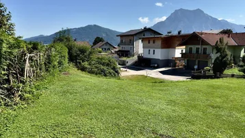 Grüne Wiese mit Blick auf umliegende Häuser und majestätische Berge im Hintergrund unter blauem Himmel.