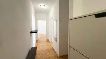 Heller Flur mit Holzboden und einfacher Einrichtung, der zu weiteren Räumen führt.