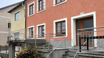 Vorderansicht eines Wohnhauses mit orangeroter Fassade, braunen Fenstern und einem Eingangsbereich mit Treppe.