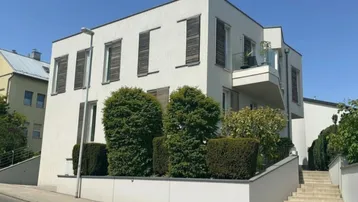 Moderne Wohnhausfassade mit mehreren Fenstern, Balkon und gepflegten Hecken im Vorgartenbereich.