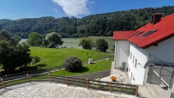 Große gepflasterte Terrasse mit Blick auf den Fluss und die grüne Landschaft, angrenzend an ein weißes Haus mit rotem Dach.