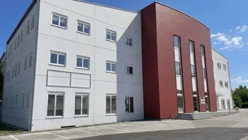 Modernes Geschäftsgebäude mit weißer Fassade und markantem roten Anbau, umgeben von Parkplätzen unter blauem Himmel.