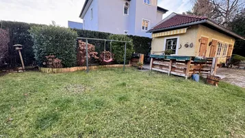 Grüner Garten mit Rasenfläche, Schaukel und einem kleinen Gartenhaus im Hintergrund.