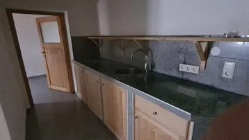 Kleine Küche mit Holzschränken, Spülbecken und dunkler Arbeitsplatte, angrenzend an einen weiteren Raum.
