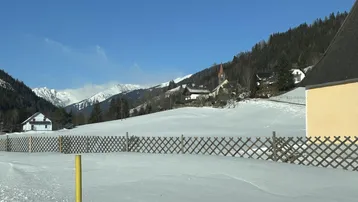 Winterliche Berglandschaft mit verschneiten Feldern, Häusern und einem Kirchturm in der Ferne unter blauem Himmel.