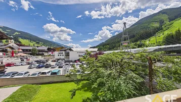 Blick vom Balkon auf die umliegende Berglandschaft, einen Parkplatz und eine Seilbahnstation.