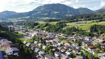 Luftaufnahme eines malerischen Dorfes in einem grünen Tal, umgeben von bewaldeten Bergen unter einem bewölkten Himmel.