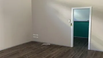 Leerer Büroraum mit Laminatboden, Klimaanlage und Zugang zu einem weiteren Zimmer mit farbiger Wand.