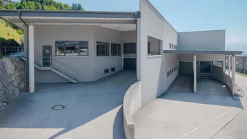 Modernes Geschäftsgebäude mit Zufahrt, überdachtem Eingangsbereich und einer geschwungenen Betonmauer.
