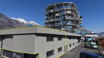 Modernes Geschäftsgebäude mit grauer Fassade im Vordergrund und einem auffälligen Glasbau im Hintergrund, umgeben von Bergen.