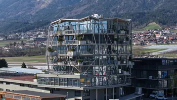 Modernes Gebäude mit markanter Glasfassade und Stahlkonstruktion, umgeben von einer Berglandschaft und städtischer Bebauung.