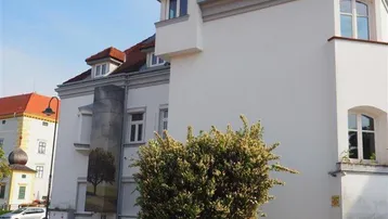 Weißes Gebäude mit rotem Ziegeldach und einem kleinen Erker, umgeben von Grünflächen.