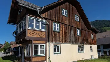 Traditionelles Holzhaus mit weiß-grünen Fenstern und heller Fassade in ländlicher Umgebung.