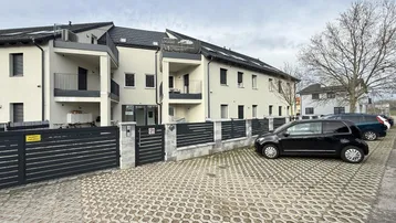 Moderne Wohnanlage mit Balkonen, Zaun und Parkplätzen unter bewölktem Himmel.